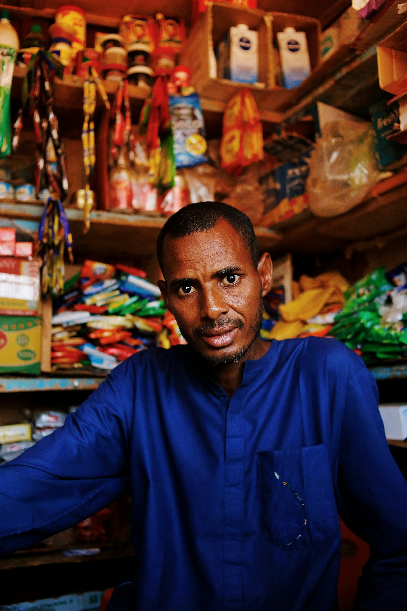 Shopkeeper in a blue shirt standing in his stocked shop